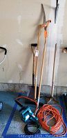 Photo showing all garden and yard tools including two electric chain saws, tree trimmer, tamper and shovel standing against a wall