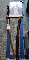 Samurai sword with black wrapped handle and sheath laid on floor with poster behind.