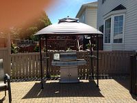 Full view of the outdoor metal frame canopy with two side shelves and missing fabric top. Shows a grill underneath the canopy.