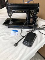 Full side view of the black Singer sewing machine showing brand name, pedal, and power cord on white surface.