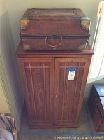 Wooden cabinet with two vertically paneled doors, warm brown finish, and woven decorative suitcase with metal hardware placed on top.