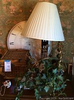 Decorative lamp with bamboo style trunk and white pleated shade alongside a round vintage-style wall clock, small figurines, and a woven wooden basket on a surface against floral wallpaper.