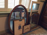 Four framed mirrors and artwork leaning against wall: oval wooden framed mirror, golden rectangular mirror with chip, crest artwork, white framed glass artwork, plus two framed art pieces on floor