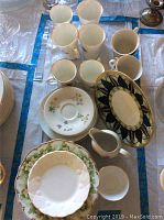 Overview photo showing multiple white mugs, floral saucers, oval plates, and a small creamer with floral decoration.