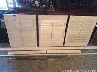 Three white vinyl window shutters with horizontal slats placed vertically alongside white framing pieces and a metal tension rod on a carpeted floor.
