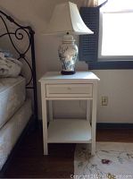 Full view of white wooden nightstand next to bed with Asian-inspired ceramic lamp on top