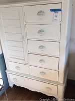 Full front view of cream colored wooden armoire dresser with six small drawers, a wide bottom drawer, and left cabinet door with cup drawer pulls.