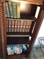 Bookshelf with three shelves containing books. Top shelf has three large hardcover history books. Middle shelf contains The Standard History of the World volumes 1-10 in brown hardcover with gold embossing. Bottom shelf has smaller paperback and hardcover books in a variety of titles and colors.