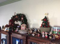 Full view of assorted Christmas ornaments, wreath, mugs, plates, and figurines displayed on wooden furniture.