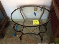 Round glass top end table with curved dark brown iridescent metal framing and a geometric lower glass shelf, positioned against a wall on a carpeted floor