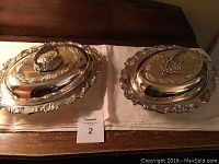 Two oval silver plate serving dishes with lids placed side by side on a white cloth over a wooden surface