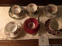 Full view of 6 assorted china tea cups with matching saucers placed in 2 rows on white cloth, showing detailed patterns and colors.