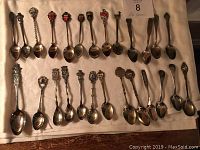 Full layout of 26 tea spoons laid out in rows on fabric surface showing varied designs and sizes, mostly silver with some showing tarnish.