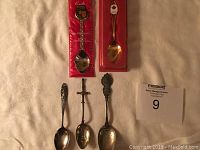 Full lot showing six tea spoons, two in original packaging, one with red and white maple leaf design, a sword-shaped handle spoon, and others with intricate detailing.