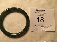 Top view of a round green jade bangle with natural stone color variations and polished finish, placed on light fabric next to a MaxSold auction card labeled 'JADE BANGLE'.
