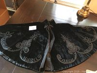 Black velvet cape laid flat showing the detailed beaded applique design and satin trim along the hem, with satin tie closure at neck.