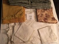 Top view of various white embroidered napkins or linens, mustard yellow fabric with fringe, and beige floral cloth.
