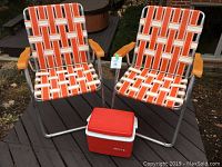 Two vintage folding lawn chairs with woven webbing in orange, white, and brown colors on a deck with wooden arm rests and metal frames. A small red and white plastic cooler is placed between the chairs.