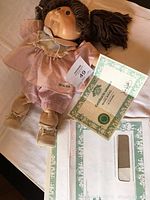 Cabbage Patch doll laying on surface next to birth certificate and adoption certificate documents, showing signed papers and doll outfit.