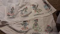 Close-up of embroidered table linens showing days Monday through Friday with colorful character designs