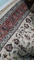 Photo showing a section of the area rug with detailed floral and leaf patterns in white, red, blue, beige, and brown tones and the red border with ornate designs.