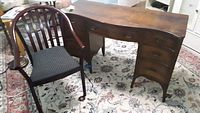 Desk with curved front and five drawers, showing scratches and one partially fixed leg, placed on a floral rug.