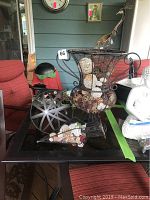 Photo showing tall metal wire vase with two compartments filled with shells, pinecones, and other decorative items. Also visible is a large decorative metal ball and a smaller wire basket on a glass table with red chairs in the background.