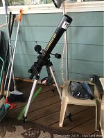 Wide view of Bushnell Voyager telescope set up on tripod on wooden floor beside various garden tools and a plastic stool.