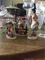 Three Hummel figurines displayed on a wooden surface with glassware in the background.