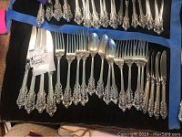 Multiple flatware pieces laid on black cloth with blue tape borders, showing entire set of knives, forks and spoons