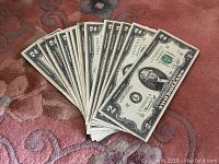 Fifteen US two-dollar bills spread out on a carpet surface, front side with serial numbers and Thomas Jefferson portrait visible.