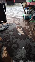 Outdoor rug showing large floral and leaf patterns in brown, beige, and cream colors, laid out on floor with furniture around.