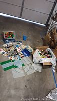 Wide shot showing a box filled with assorted LEGO bricks, several road base plates laid out on the floor, and multiple Robotix instruction booklets spread around.