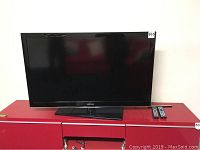 Front view of Samsung television placed on red TV stand with two remote controls.