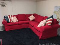 Red Ikea corner sofa with five pillows arranged on it in a room with concrete walls and black carpet.