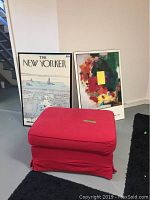 Red fabric ottoman with fabric cover, rectangular with rounded edges, placed on floor near two framed pieces of wall art.