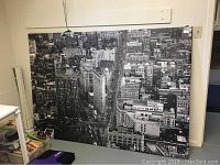 Large black and white aerial cityscape canvas wall art leaning against a wall indoors, showing detailed city buildings and streets.