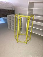 Yellow hexagon metal side table with glass top, shown next to white shelving units and bed frame