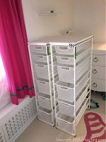 Two white Elfa metal drawer units side by side with six mesh drawers each, placed next to a window with pink curtains.