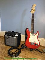 Full view of red Squier Mini electric guitar on stand next to black Fender amplifier and coiled cable on hardwood floor.