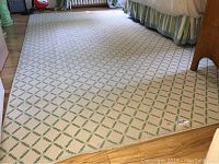 Photo shows the full length view of the large area rug placed on a wooden floor next to furniture, displaying the repeating diamond pattern with green leaves on a light background.