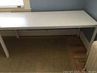 Front view of large white rectangular table/desk showing smooth flat top and four legs placed on a rug and hardwood floor.