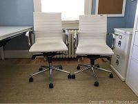 Two white office chairs placed side by side in front of a radiator and window, showing front view of seats and chrome armrests.
