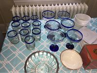 Photo showing clear drinking glasses with cobalt blue rims, some large glasses, white bowls and stacks of white plates on light blue patterned tablecloth.