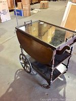 Angled side view of the vintage beverage cart showing large wheels, glass top, and lower shelf
