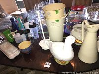 View of collection including ceramic pitchers, buckets, duck-shaped ceramic container, glassware, and other ceramics on a wooden table.