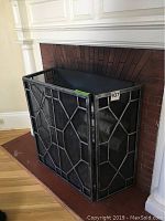 Fireplace grate placed inside a fireplace opening, showing front and side geometric patterned metal panels.