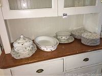 Overview of tureen with lid and undertray at left, two oval bowls, round bowl on plate, stack of plates at right on wooden surface under shelf