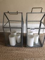 Wide shot showing two metal-framed glass lanterns side by side on carpeted floor with white pillar candles inside each lantern.