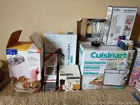 Photo showing all boxed and some loose kitchen appliances and tools including Cuisinart breadmaker, Oster ice cream maker, Braun chopper, and others